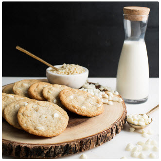 A glass of milk next to a batch of white chocolate macadamia cookies on a wooden tray.